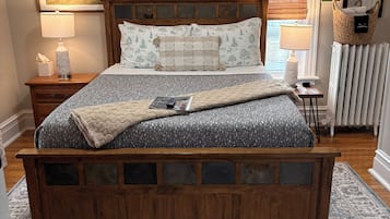 Een slaapkamer met een houten bed, een nachtkastje, een venster met zonwering, een radiator en een kamerkleed met dessin.