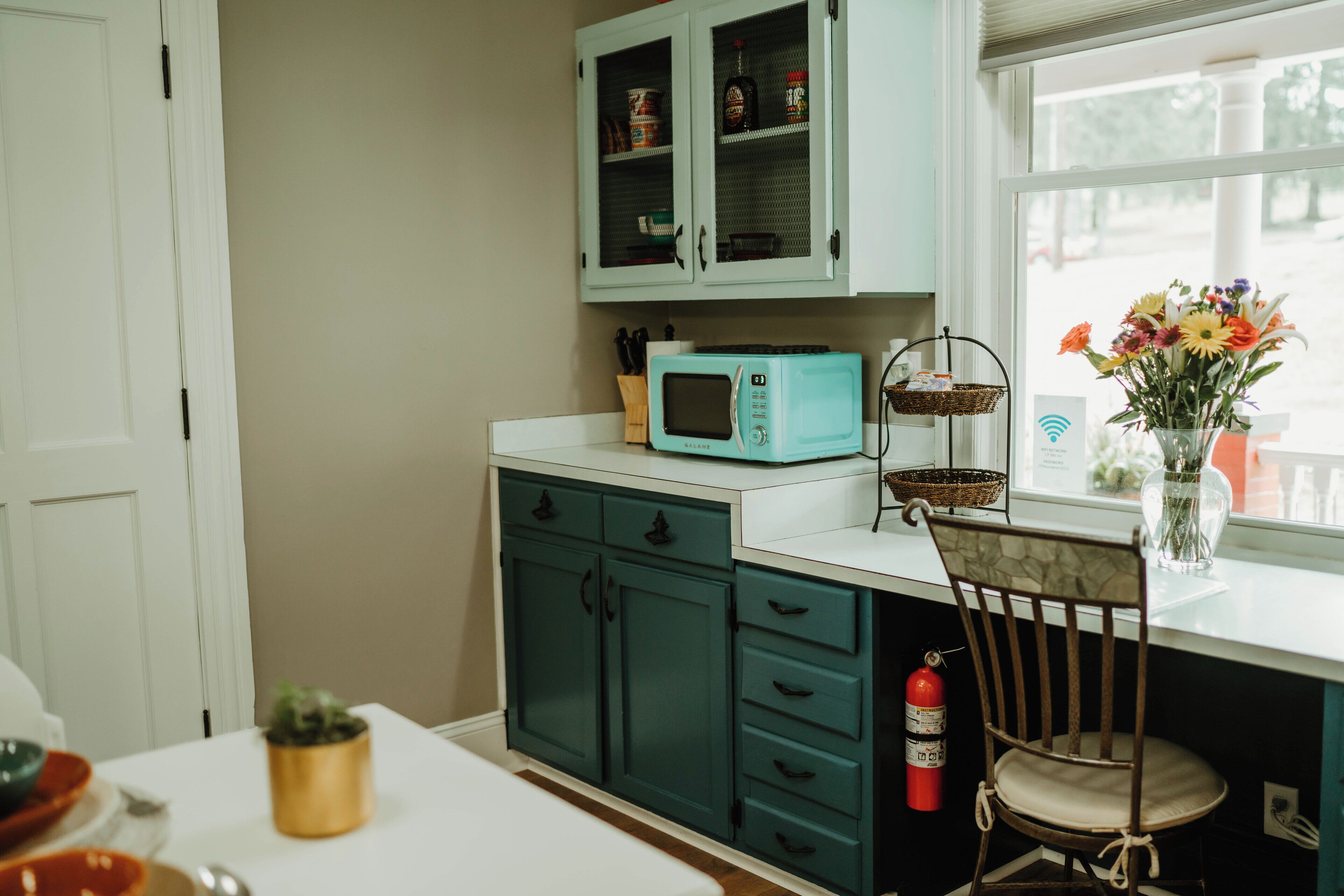 Una cocina con armarios color turquesa, un microondas, una silla, un extintor y una ventana con flores.