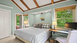 Une chambre à coucher avec un lit, un bureau avec un ordinateur, une chaise, une fenêtre donnant sur des arbres et une porte.