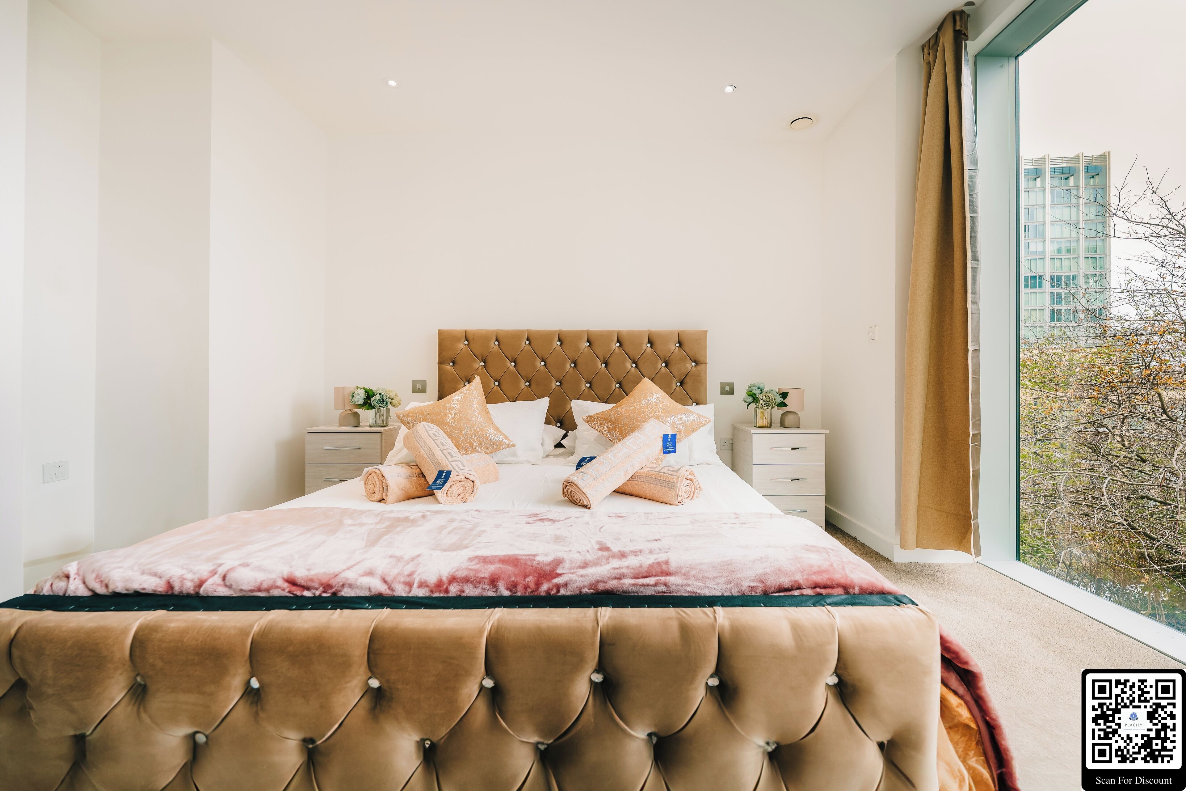 Ein modernes Schlafzimmer mit einer gepolsterten Kopfteile, einem großen Bett mit einer rosa und weißen Bettdecke und Blick auf eine Stadtlandschaft durch ein großes Fenster.