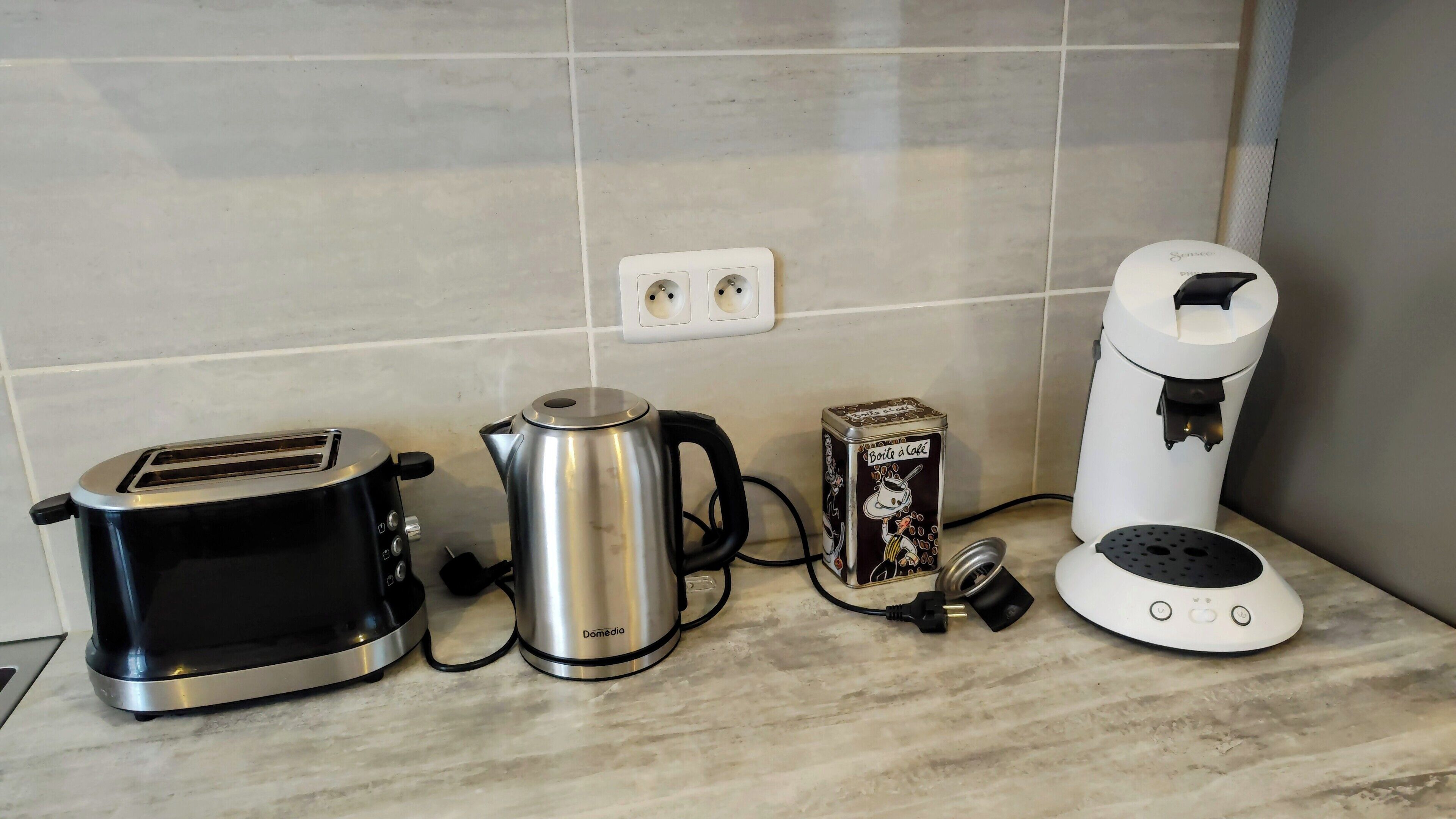 Un plan de travail de cuisine avec un grille-pain, une bouilloire, une machine à café et une petite boîte en métal.