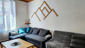 Una sala de estar moderna con un sofá gris, una obra de arte de madera en la pared, una mesita con una planta y una ventana con cortinas geométricas.