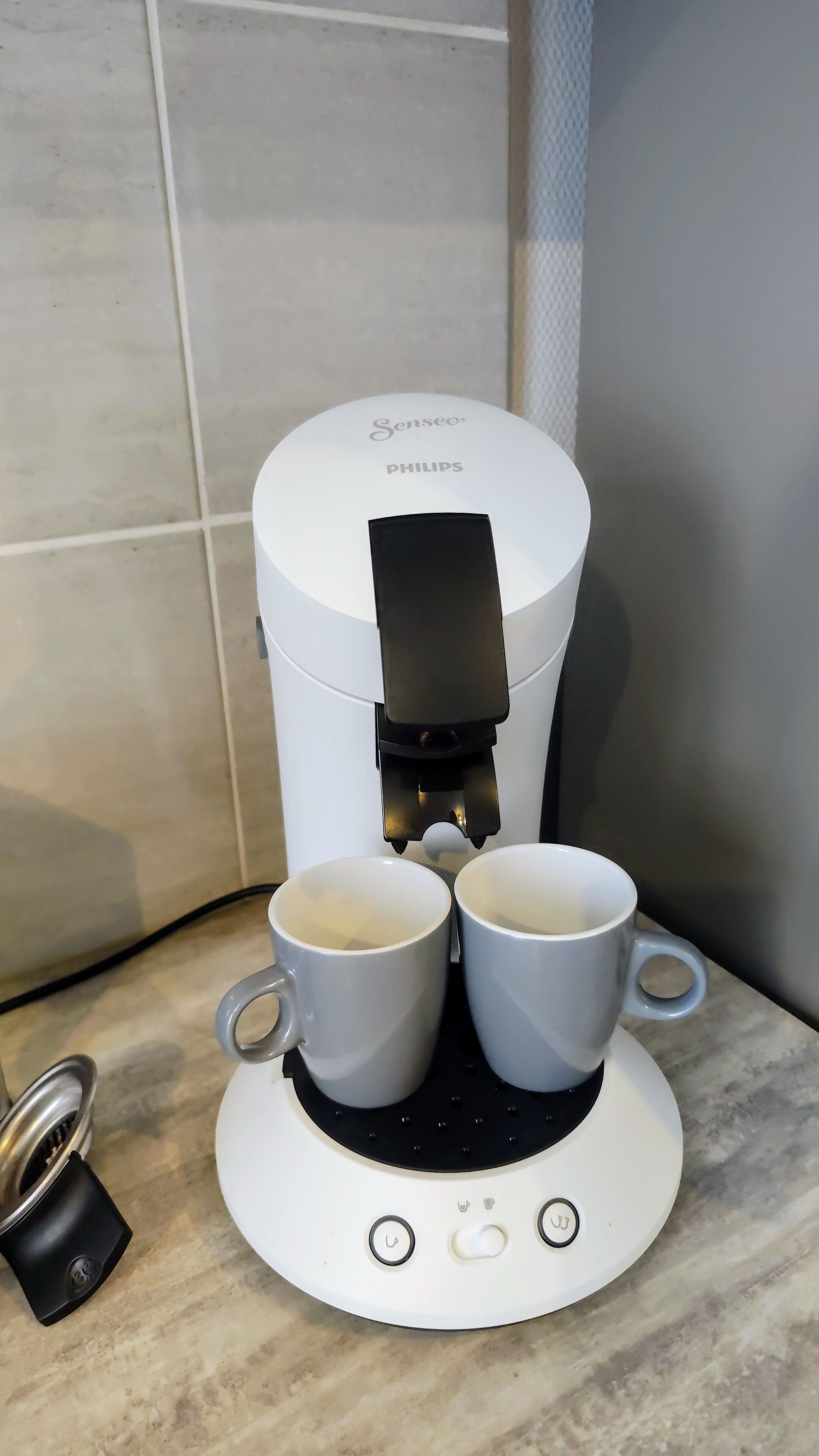 A Senseo Philips coffee maker with two white cups on its base, a power cord, and a small black device on the counter.