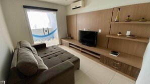 A modern living room with a sofa, TV, wooden entertainment unit, and a view of a blue sky. - Casa com Piscina - Quartos Privativos (Sao Raimundo Nonato)