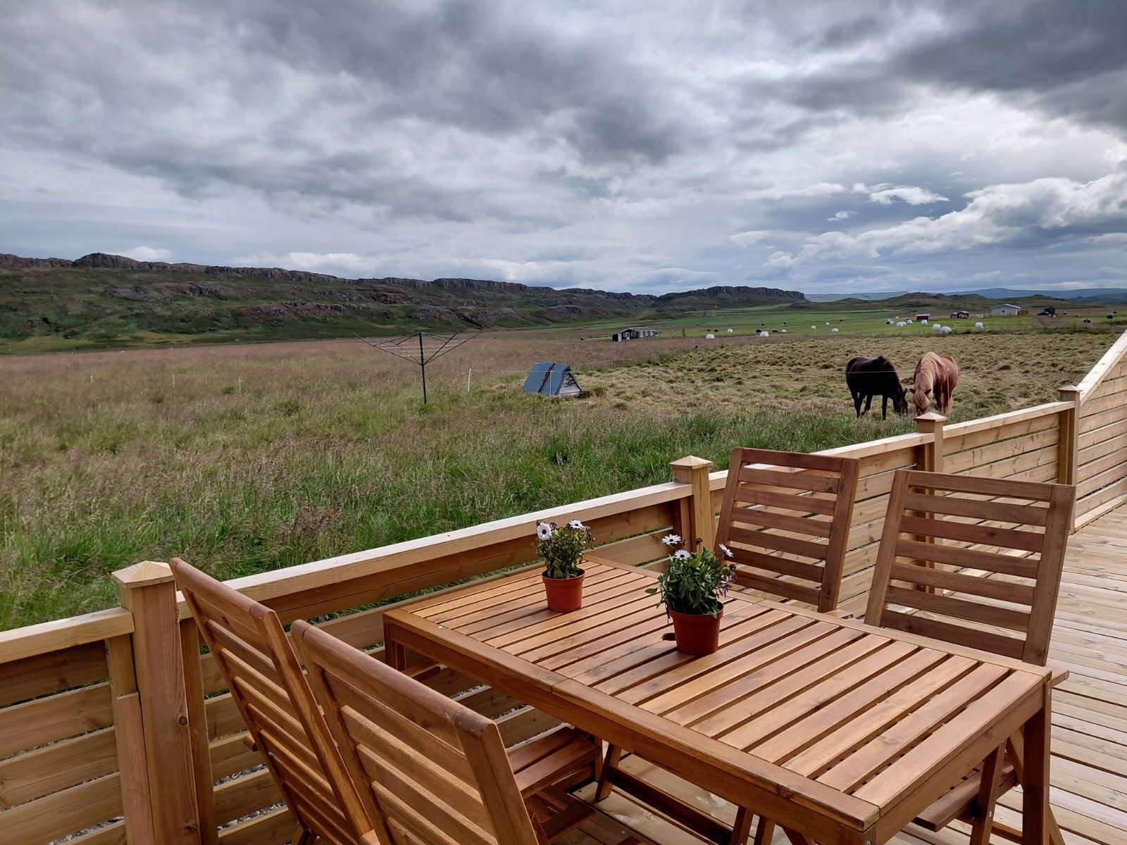 Um deck de madeira com mesa e cadeiras, com vista para uma paisagem rural onde animais pastam e há uma tenda.