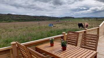 Um deck de madeira com mesa e cadeiras, com vista para uma paisagem rural onde animais pastam e há uma tenda.