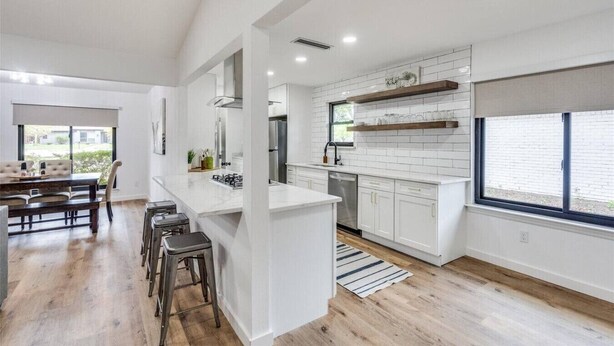 A modern kitchen with white cabinetry, a central island, and a dining area with a wooden table and bar stools. - LUX Lounge | 3200+ SQ FT - Heated Pool & Jacuzzi (Richardson)