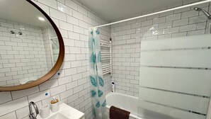 A bathroom with white tiled walls, a white sink with a wooden countertop, a mirror with a wooden frame, a glass shower enclosure, and a towel rack with a brown towel. - Rooms Borrell (Barcelona)