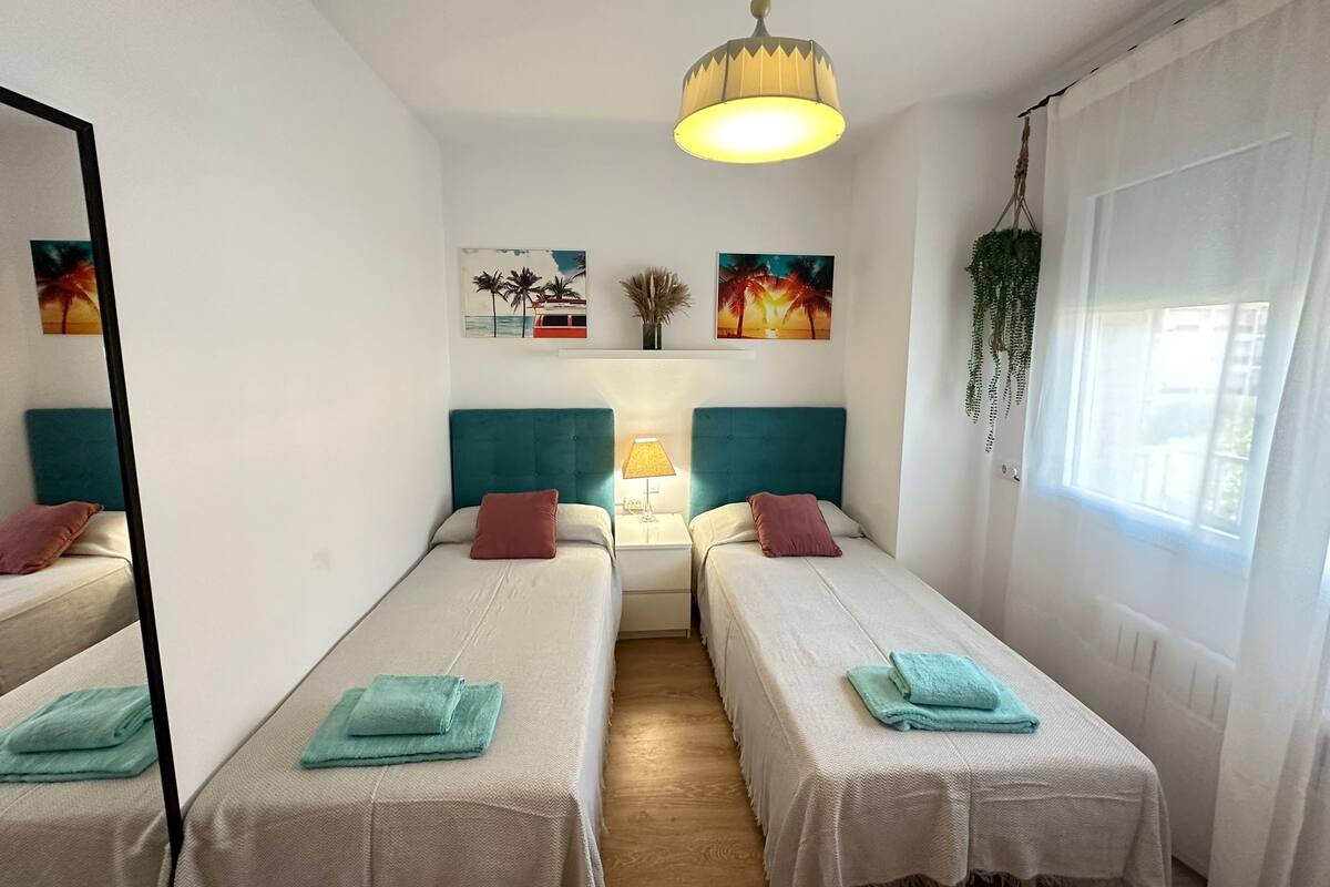 Una habitación con dos camas individuales, cada una con cabecero color aguamarina y ropa de cama blanca. Hay dos lámparas de noche, una planta colgante y una obra de arte en la pared.
