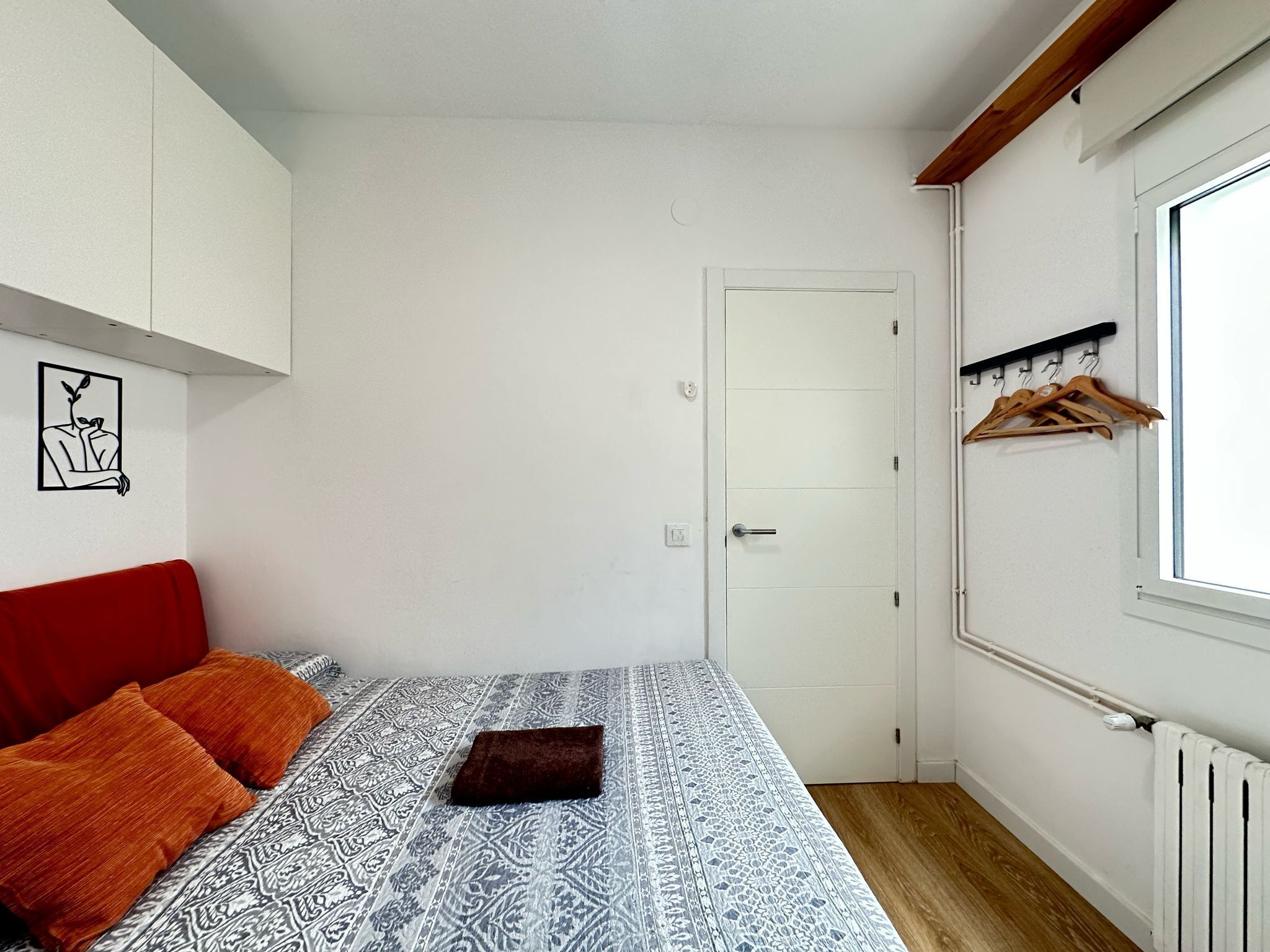 A small, neatly arranged bedroom with a bed, a red couch, a patterned bedspread, a door, a window with a wooden shutter, and a wall-mounted coat rack.