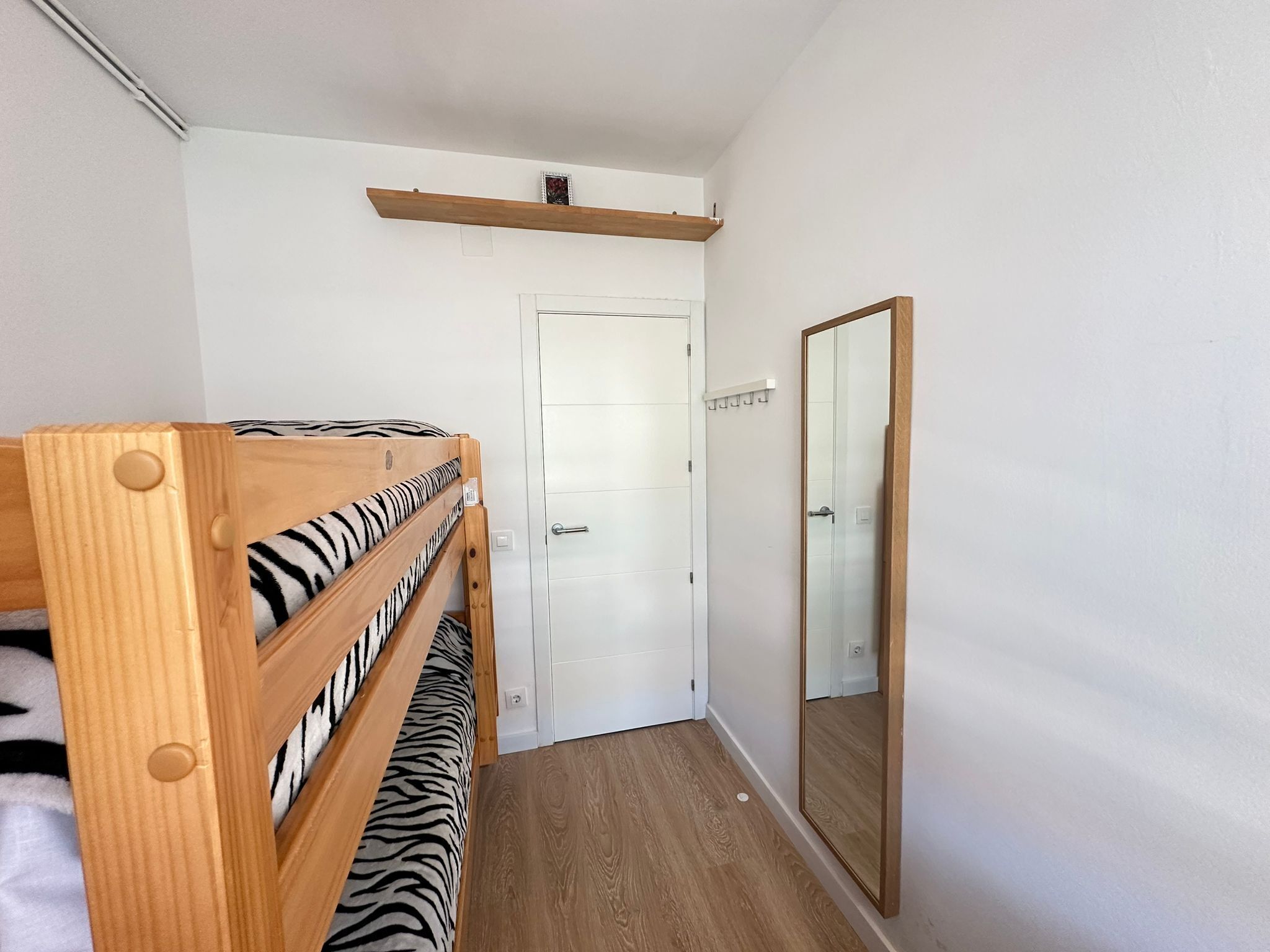 A narrow hallway with a wooden bunk bed with zebra-patterned bedding, a white door, and a large mirror on the right wall.