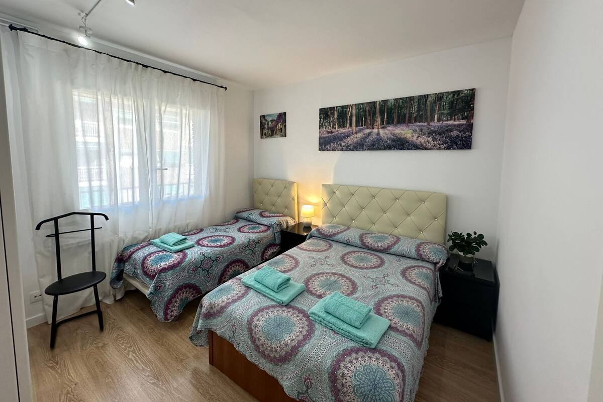 Un dormitorio con dos camas, una silla, una mesita de noche, una lámpara y un cuadro en la pared.