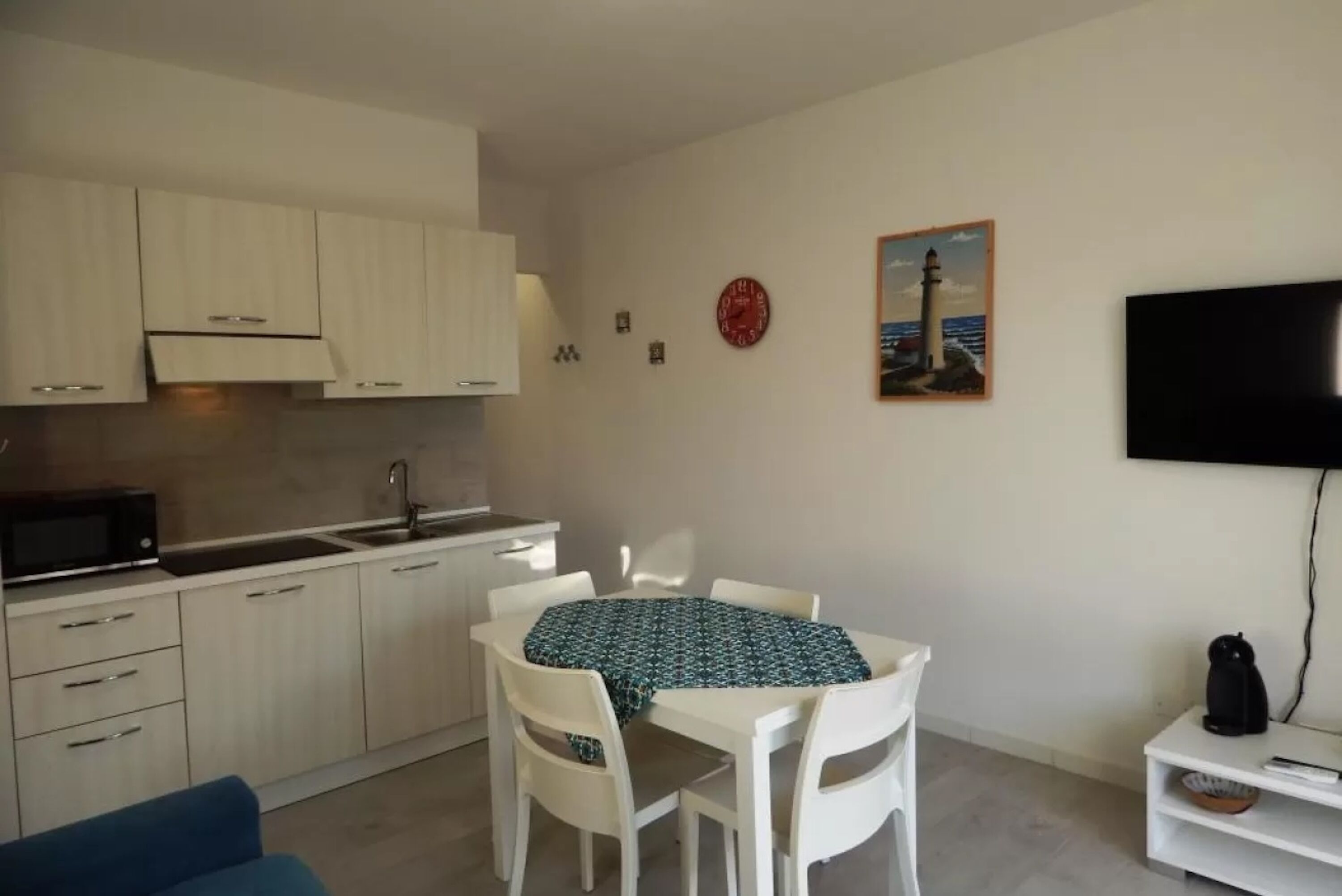 Une cuisine compacte avec des meubles blancs, un four à micro-ondes et un évier. On y trouve également un coin repas avec une table et des chaises, une horloge et un tableau représentant un phare.