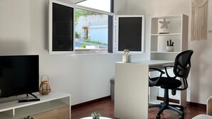 Una sala de estar moderna con televisión, un escritorio y una silla. Se ve una ventana con vistas a zonas verdes y un edificio.