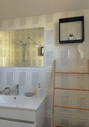 Een badkamer met een grote spiegel, een douche, een wastafel met kraan, een zeepdispenser en een kleine plant in een pot aan de muur.