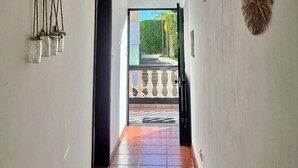 Un pasillo con paredes blancas, techo de madera y suelo de baldosas. Hay una puerta que da al exterior, un adorno en la pared y un macramé colgante.