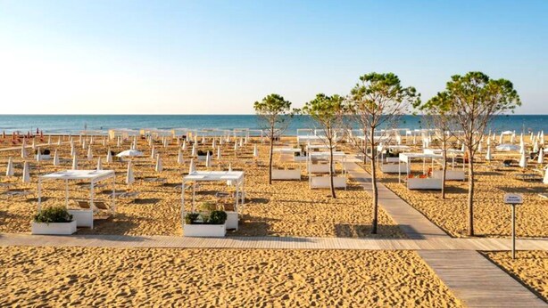 A beach with white lounge chairs and tables arranged in rows, a wooden walkway, and a few trees. - Stylish flat for 7 people - Beahost (Bibione)