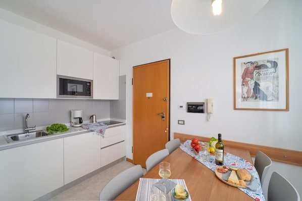 Una cocina moderna con armarios blancos, un microondas y una cafetera. Hay una mesa de comedor con una botella de vino, una copa y un poco de pan. Un cuadro enmarcado cuelga en la pared.