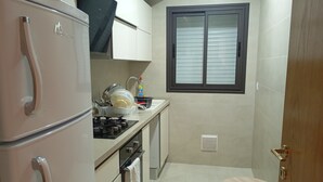 A compact kitchen with a refrigerator, stove, and sink. There is a window with blinds, a small stool, and a vent on the wall. - VIP Tunisia  (Halq al Wadi)