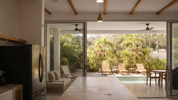A modern kitchen with a large island, a dining area with outdoor seating, and a view of a lush garden and pool.