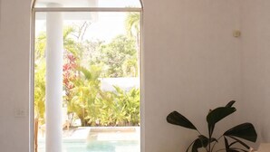 A room with a white sofa, a round ottoman, a potted plant, a wooden ceiling beam, and a glass door leading to an outdoor pool area.