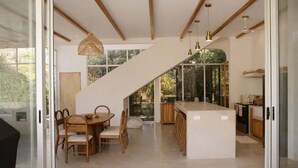 A modern kitchen with a central island, wooden dining table, and chairs, with a staircase leading to a balcony.