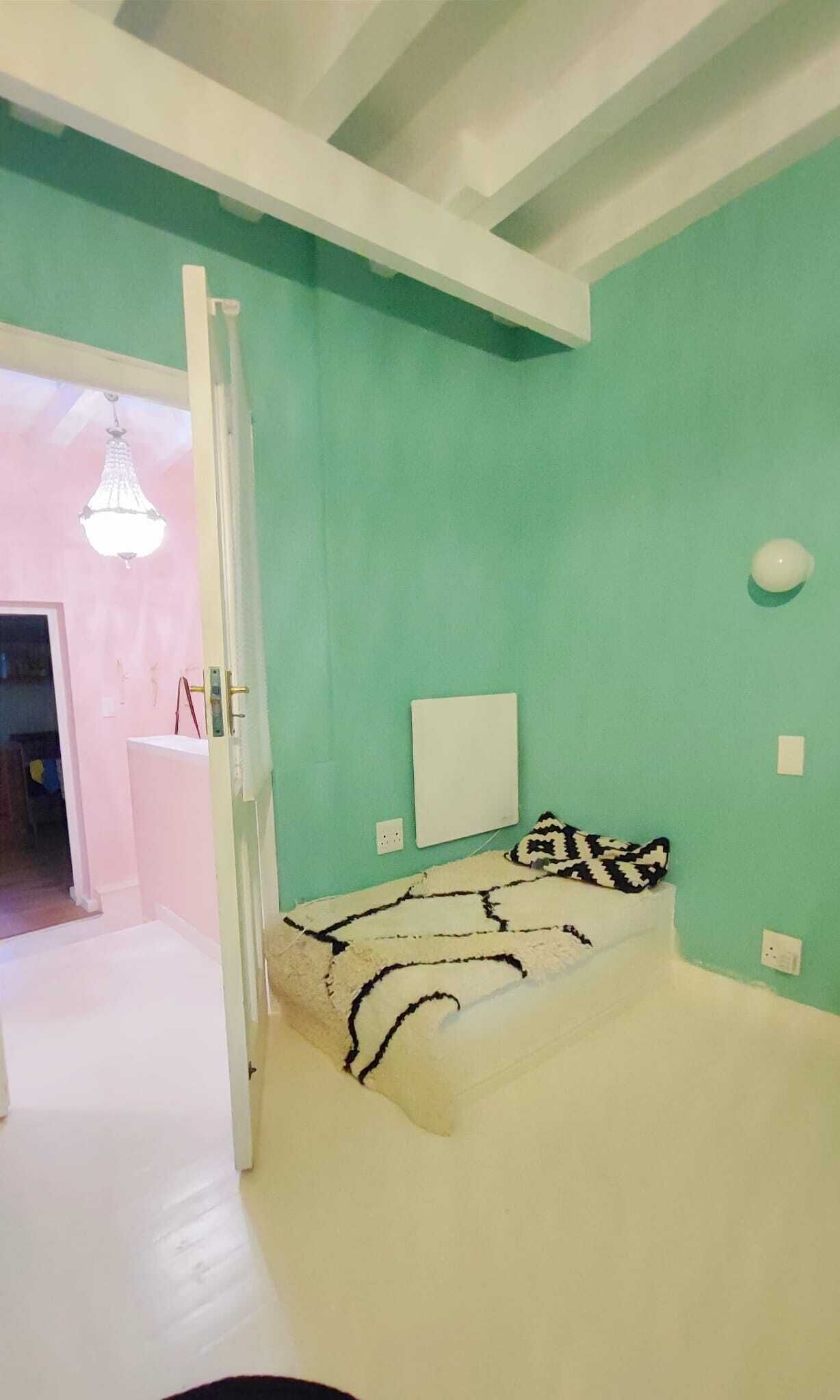 A room with teal walls, a white ceiling, and a bed with a patterned blanket. There is a door leading to another room, a white wall-mounted light fixture, and a small white object on the wall.