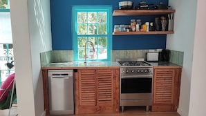A compact kitchen with a dishwasher, stove, and oven. A window with a blue frame and a view of greenery is visible. - Beautiful Appartement With Balcony and Great View at Bokaap (Cape Town)