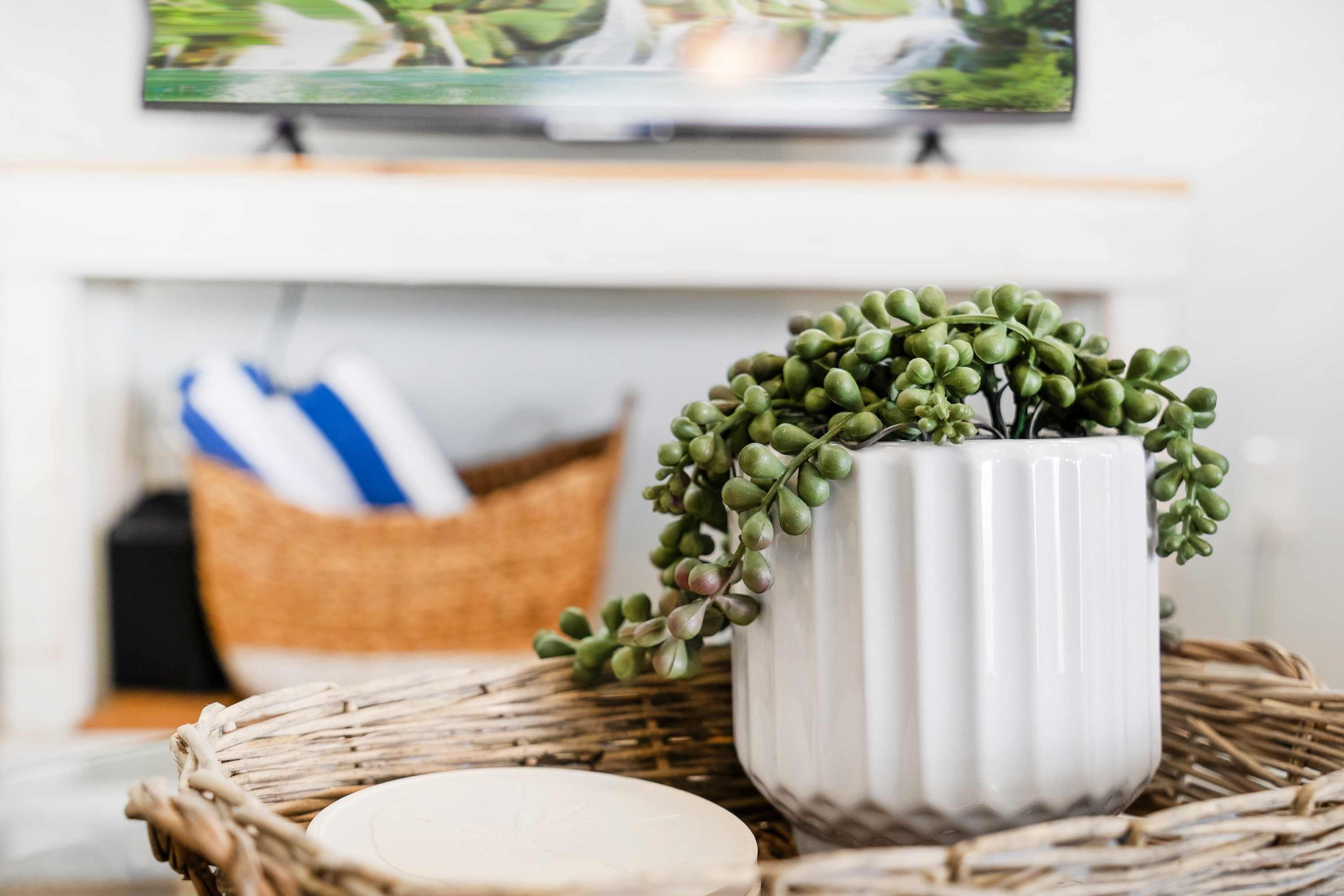 Een witte keramieken vaas met groene bladeren op een rieten mand, een witte kaars, en een blauw-wit gestreept kussen op de achtergrond.