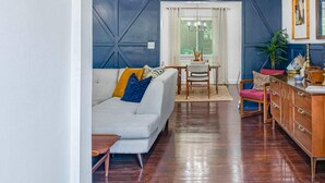 Una sala de estar con sofá, una mesa de comedor con sillas, un armario de madera y una ventana con cortinas.