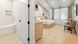 A modern hotel room with a large bed, a separate sitting area, a small kitchenette, and a bathroom with a bathtub.