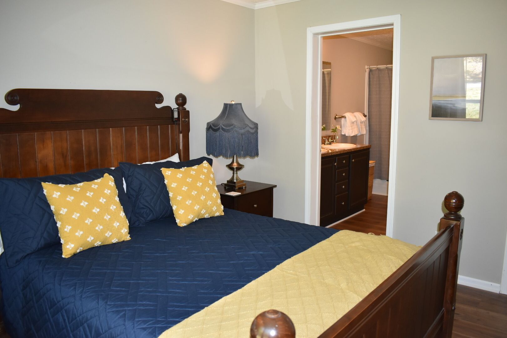 A bedroom with a wooden bed, a nightstand with a lamp, and a bathroom visible through an open door.