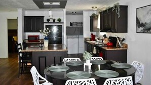 A modern kitchen with dark cabinetry, a central island with seating, and a dining area with white chairs. - Waterfront LKN Hideaway Relax & Recharge (Troutman)