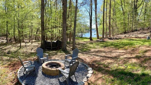 A circular seating area with a fire pit surrounded by chairs and a gravel border, set in a wooded area with a lake in the background. - Waterfront LKN Hideaway Relax & Recharge (Troutman)