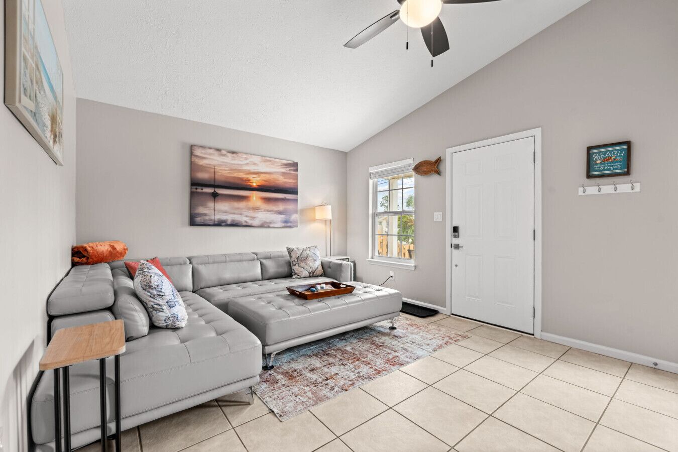 A modern living room with a grey sectional sofa, a ceiling fan, a large abstract painting, and a door with a beach-themed sign.