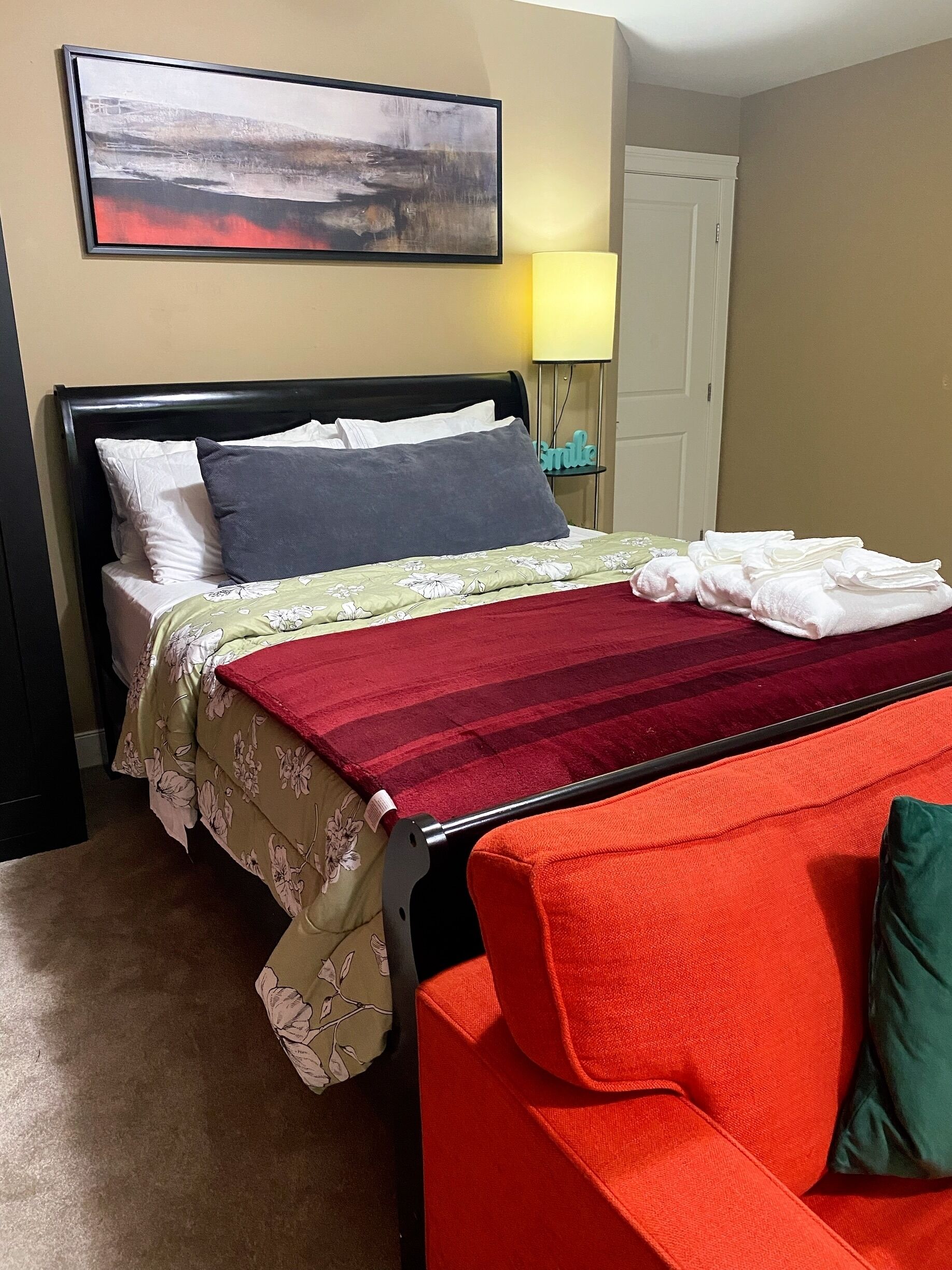 Une chambre d’hôtel avec un lit, une couverture à rayures rouges et vertes, une chaise rouge, une table de chevet avec une lampe et un tableau accroché au mur.
