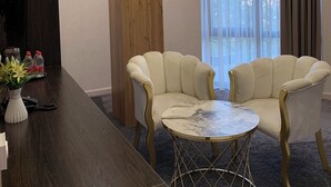 Un vestíbulo de hotel con dos sillones, una mesa de centro de mármol, una columna decorativa de madera y un amplio ventanal con cortinas.