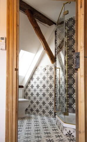 A bathroom with a sloped ceiling, a shower with a glass door, and patterned tiles. - Maison Da Silva (Oberderdingen)