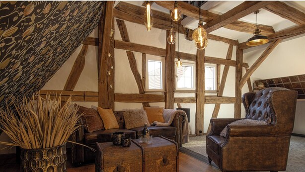 A rustic attic room with exposed wooden beams, a leather armchair, a vintage trunk, and a patterned tapestry. - Maison Da Silva (Oberderdingen)