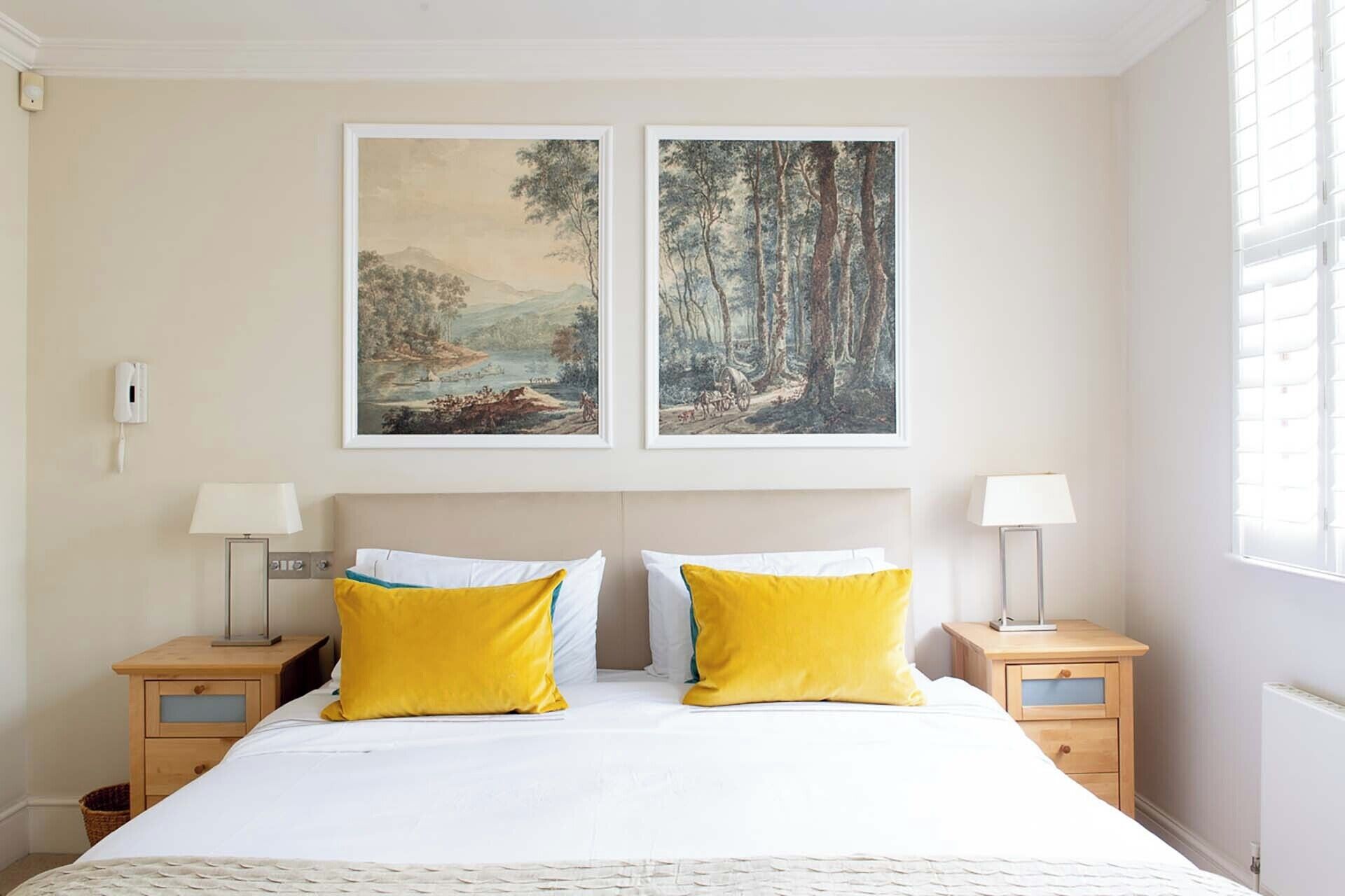 A bedroom with a large bed, two bedside tables with lamps, and two framed landscape paintings above the bed.