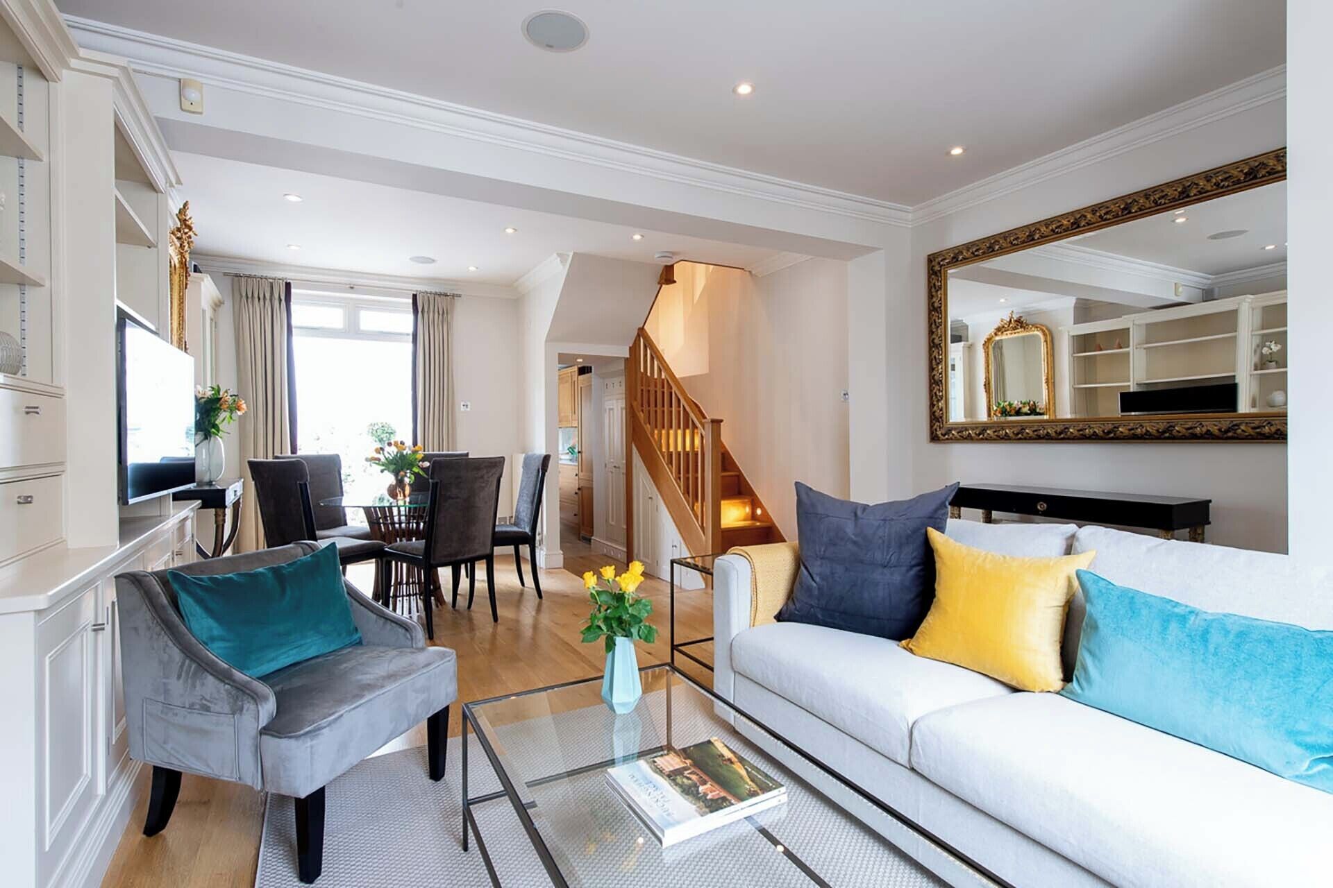 A spacious living room with a white sofa, a grey armchair, and a glass coffee table. A staircase leads to an upper floor, and a large mirror hangs on the wall.
