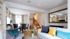 Un salón amplio con un sofá blanco, un sillón gris y una mesa de centro de vidrio. Una escalera conduce a un piso superior, y un espejo grande cuelga de la pared.