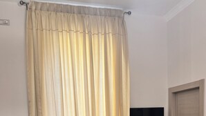Una stanza con una tenda color beige, una poltrona beige, un pavimento in legno, un televisore a schermo piatto e una porta in legno.