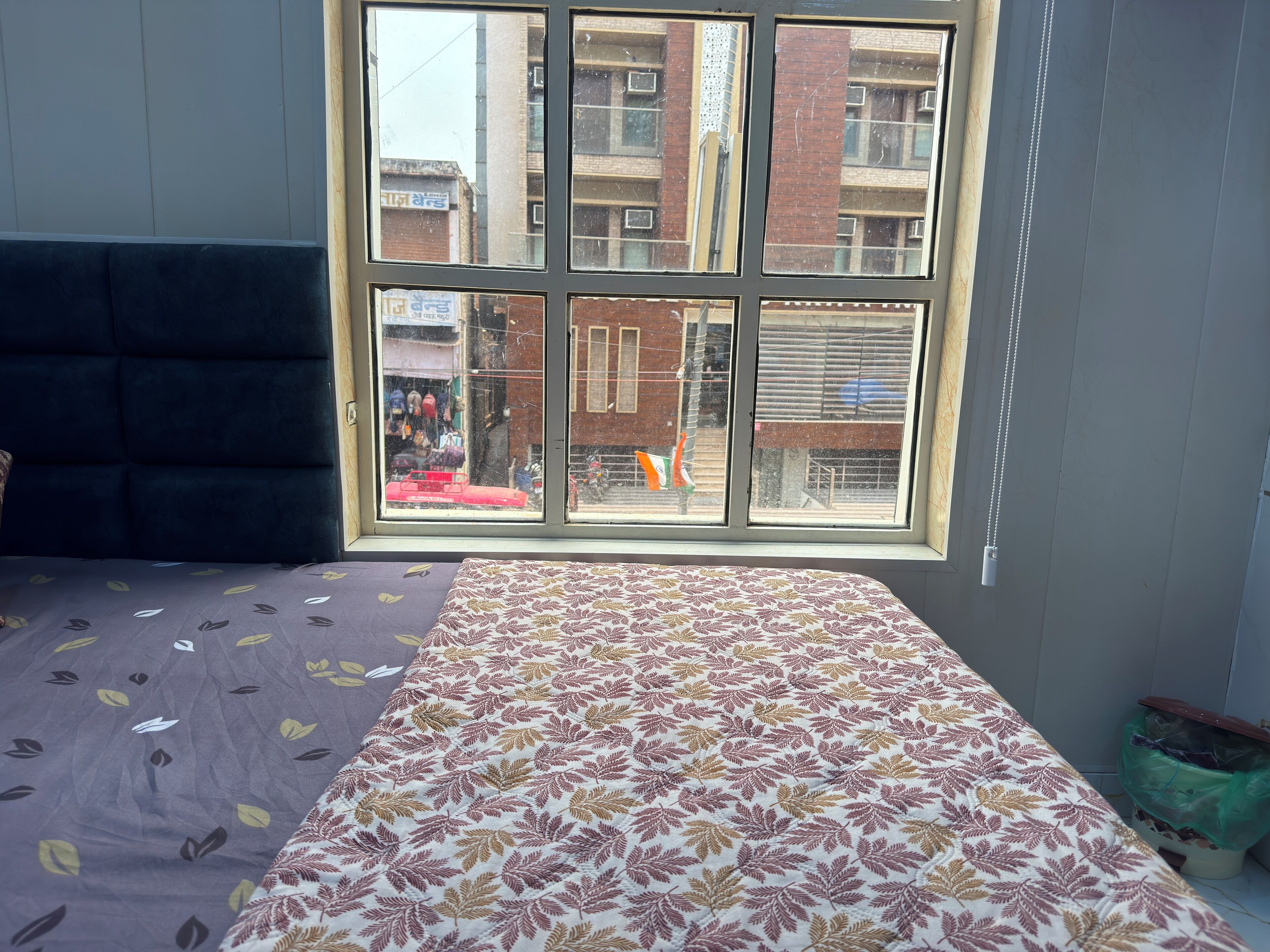 A room with a large window, a bed with a patterned bedspread, a dark blue headboard, and a green trash bin.