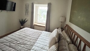 Ein Schlafzimmer mit Bett, einem Fenster mit Vorhängen, einem an der Wand montierten Fernseher, einer Nachttischlampe und einem Blumentopf.
