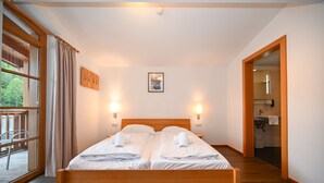 A double bed with two pillows and two towels, wooden headboard, two wall-mounted lights, a framed picture, and a view of the outdoors through a window.