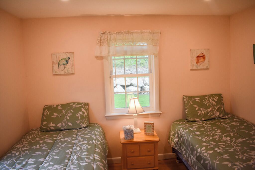 A bedroom with two beds, a window with a lace curtain, and two wall-mounted paintings.