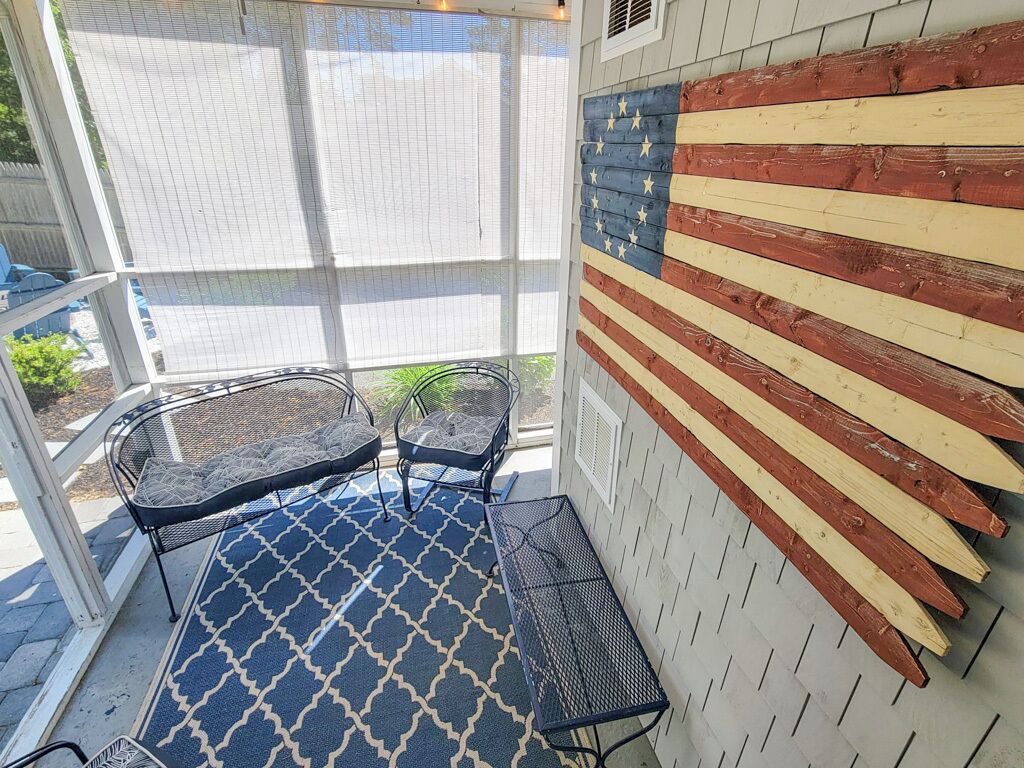 Eine Terrasse mit blauem gemustertem Teppich, Metallstühlen und einer Wand, die mit einer hölzernen amerikanischen Flagge verziert ist.