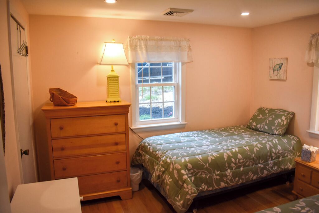 Une chambre avec une commode en bois, un lit recouvert d’une literie à motifs floraux, une fenêtre donnant sur un beau paysage et une table de chevet avec une lampe.