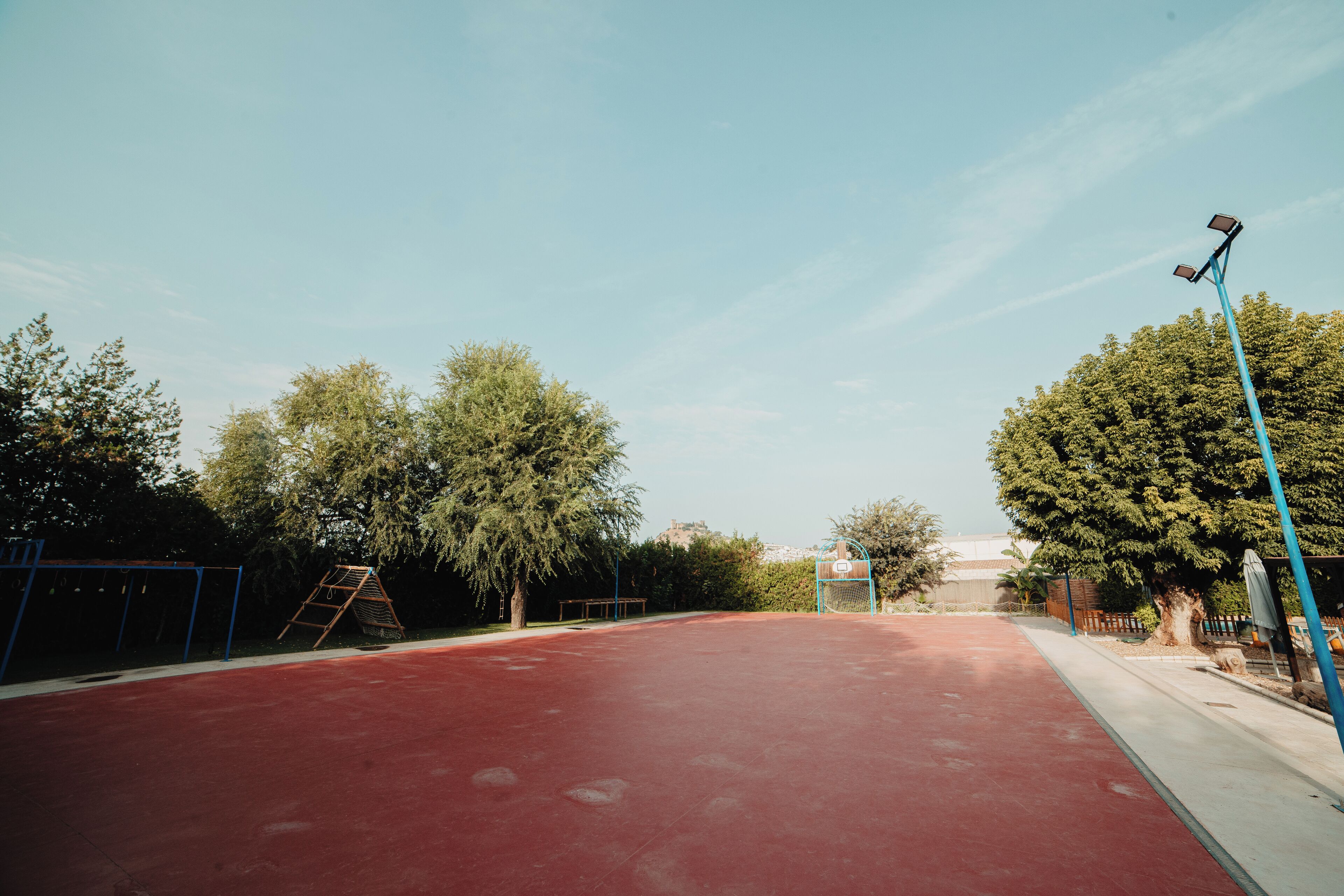 An outdoor basketball court with a red surface, surrounded by trees and a blue fence.